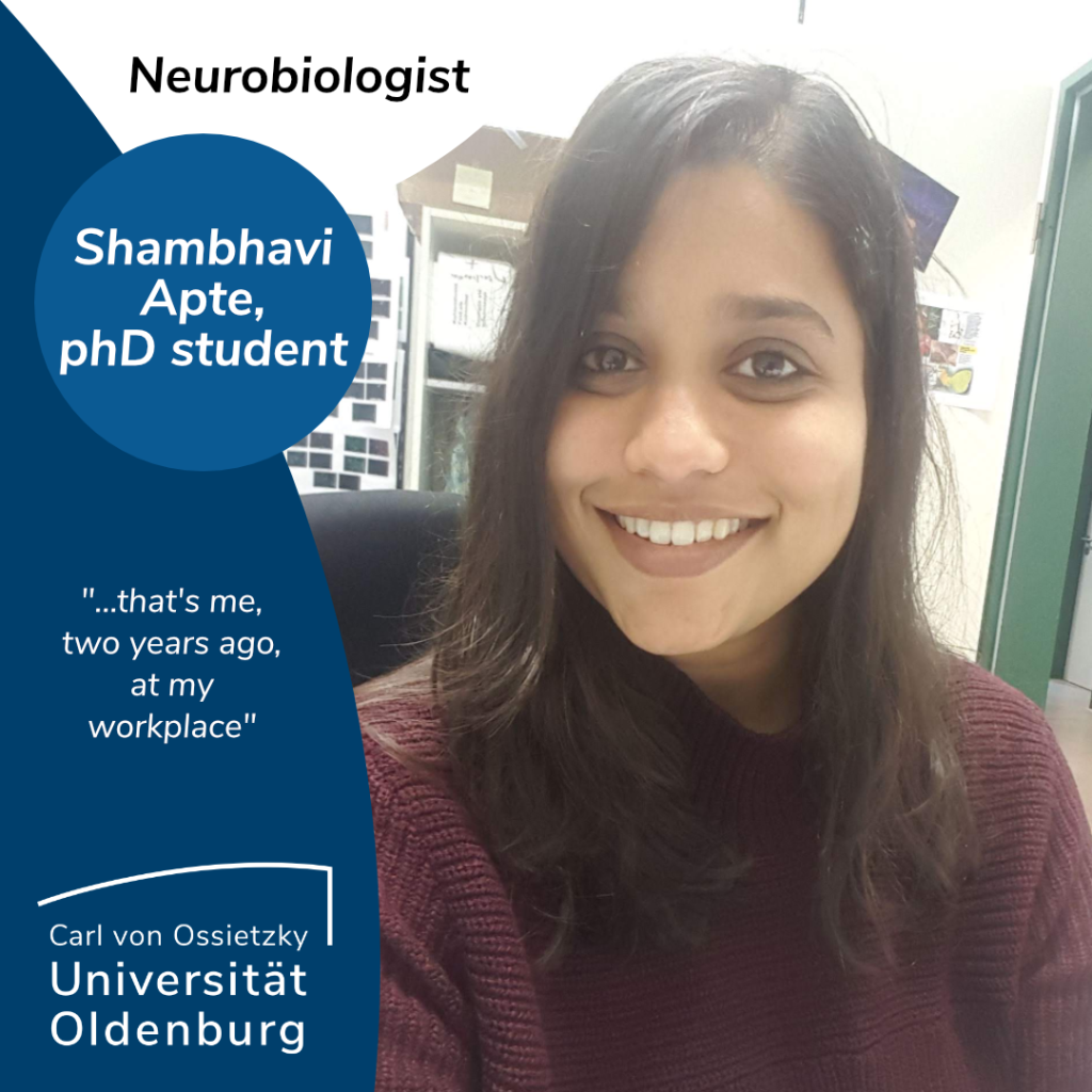 Interview with Shambhavi Apte, a phD student and neurobiologist. She says: "...that's me, two years ago, at my workplace". She, a Person of Color, wears a plum-colored sweater and has dark, slightly longer than shoulder-length hair. She is smiling at the camera and is sitting in an office. 