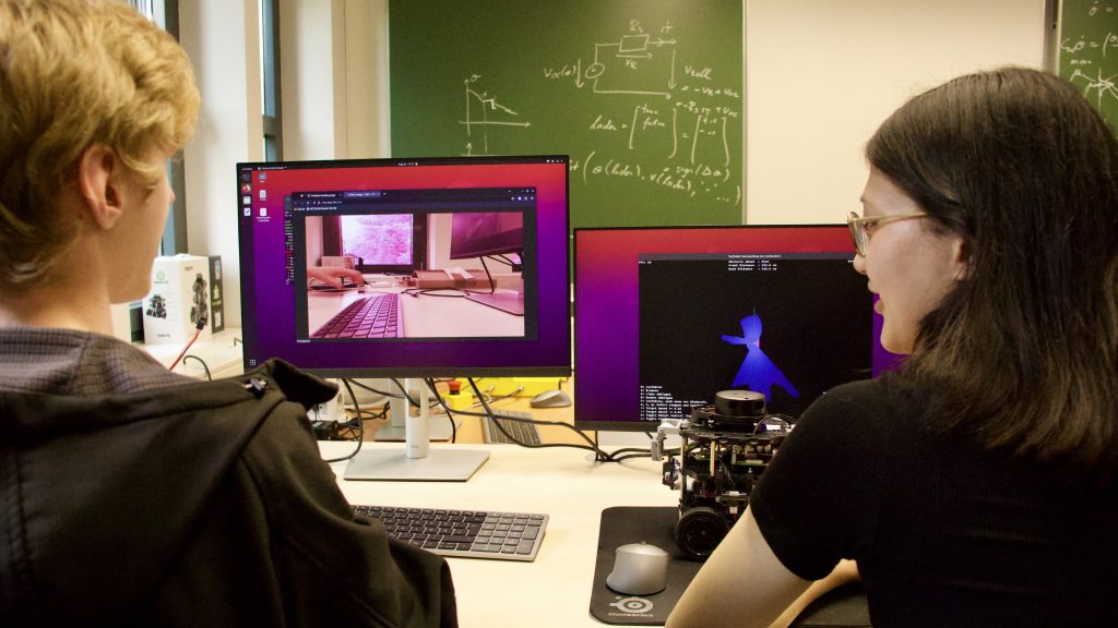 Zwei Menschen sitzen nebeneinander an einem Schreibtisch, auf dem zwei Monitore stehen. Im Hintergrund sind Schultafeln zu sehen.